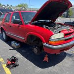 Red Chevrolet Blazer supported on jack stands with the hood open and front wheel removed during brake repair service.