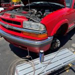 Front view of a red Chevrolet Blazer with the hood open during brake repair service, showing the front wheel removed and brake components exposed.