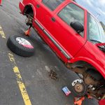 Red Chevrolet Blazer SUV raised on jack stands during rear brake repair, with wheels removed and rust visible along the rocker panels.