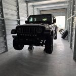 Black Jeep Wrangler supported on jack stands inside a storage unit during suspension and steering repair work, with wheels removed for service.