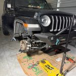 Black Jeep Wrangler lifted for front suspension repair with the front wheel removed, exposing steering and suspension components.