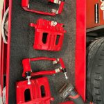 Two freshly painted red brake calipers and their mounting brackets are arranged on a black workbench mat beside a cordless impact driver.