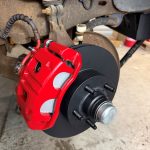 Close-up of a completed vehicle brake assembly with a new black brake rotor, freshly painted bright red brake caliper, and brake pads installed on the wheel hub.